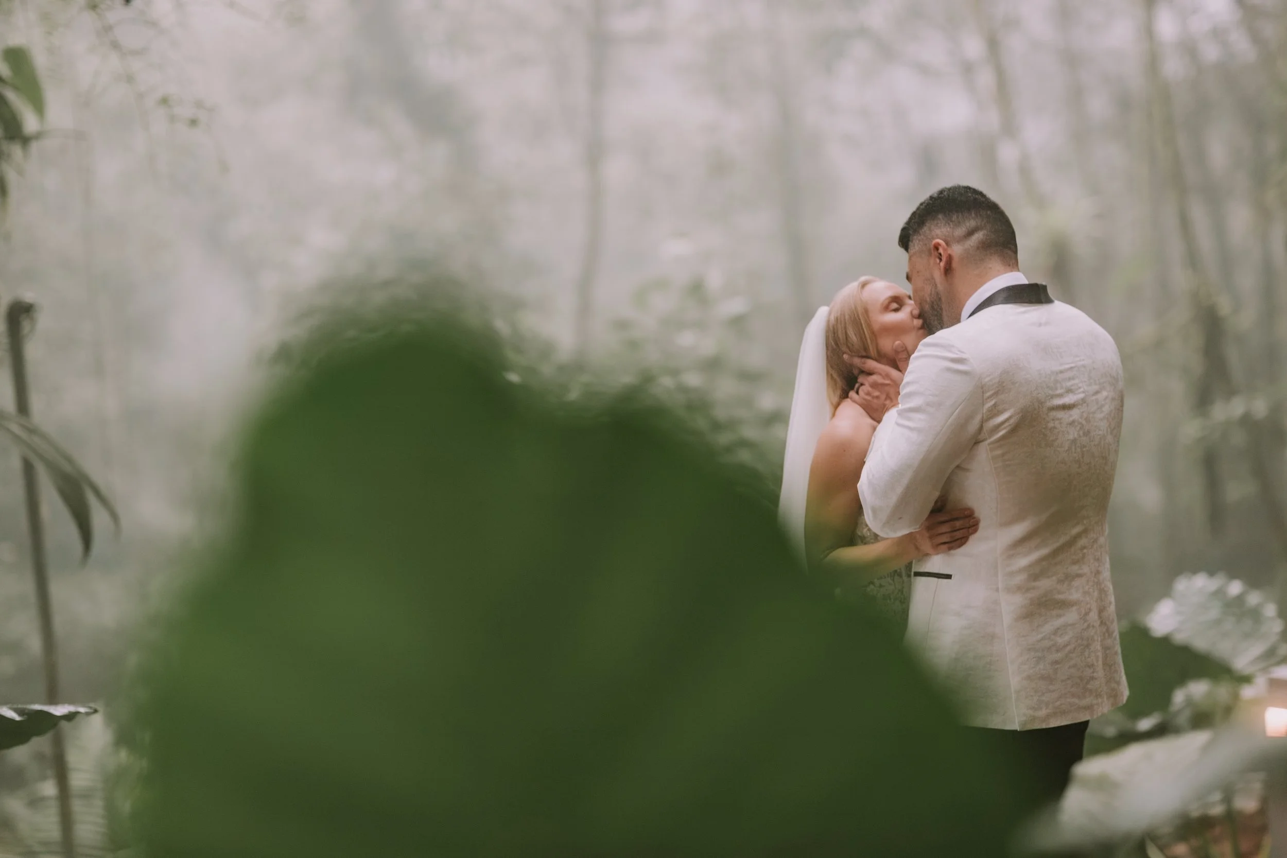 Bride with a long veil kissing her groom in a white tuxedo jacket in the misty Costa Rica cloud forest