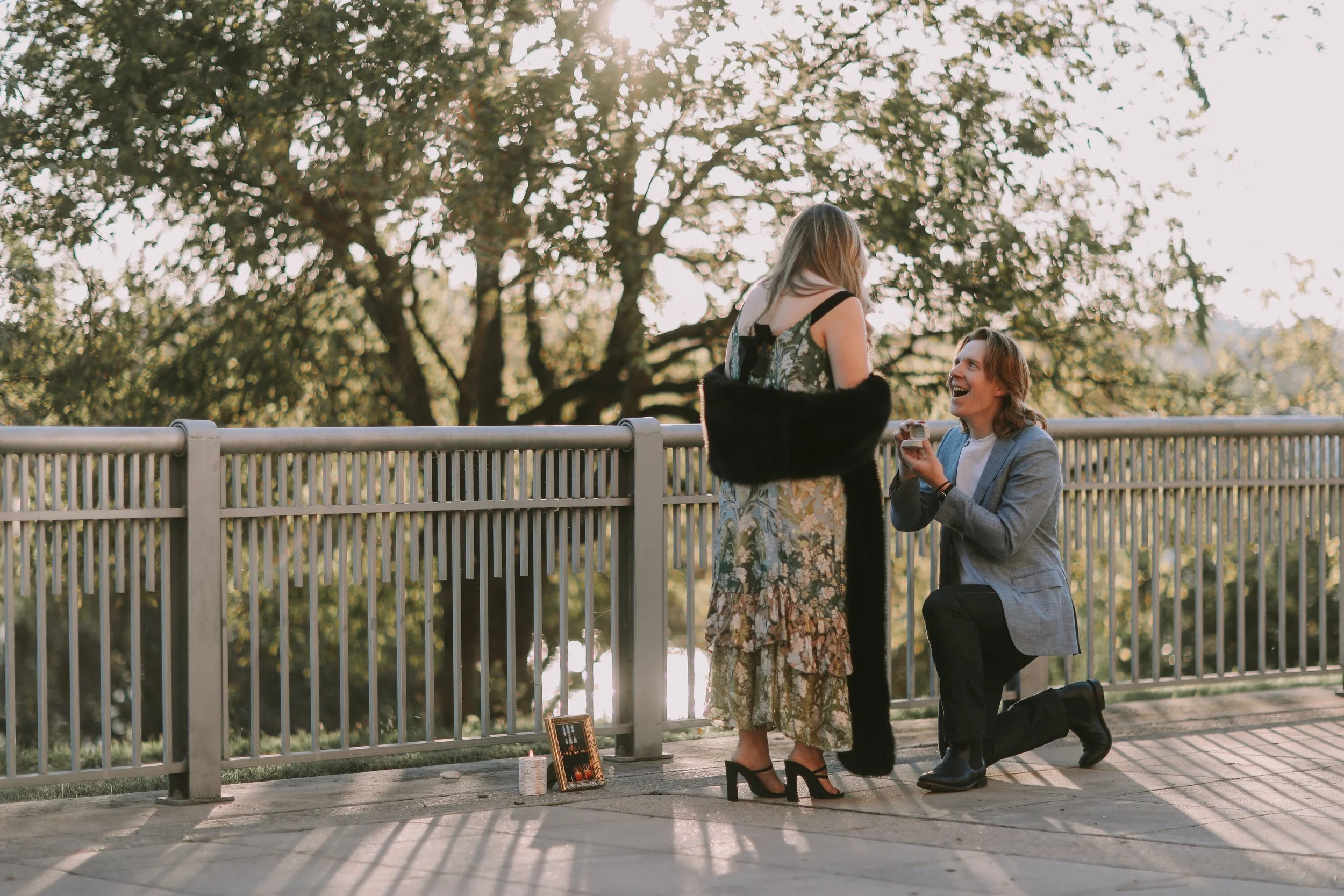 Jake kneeling with a ring as Riley, in a floral dress and black shawl, reacts on a sunlit riverside walkway in Austin