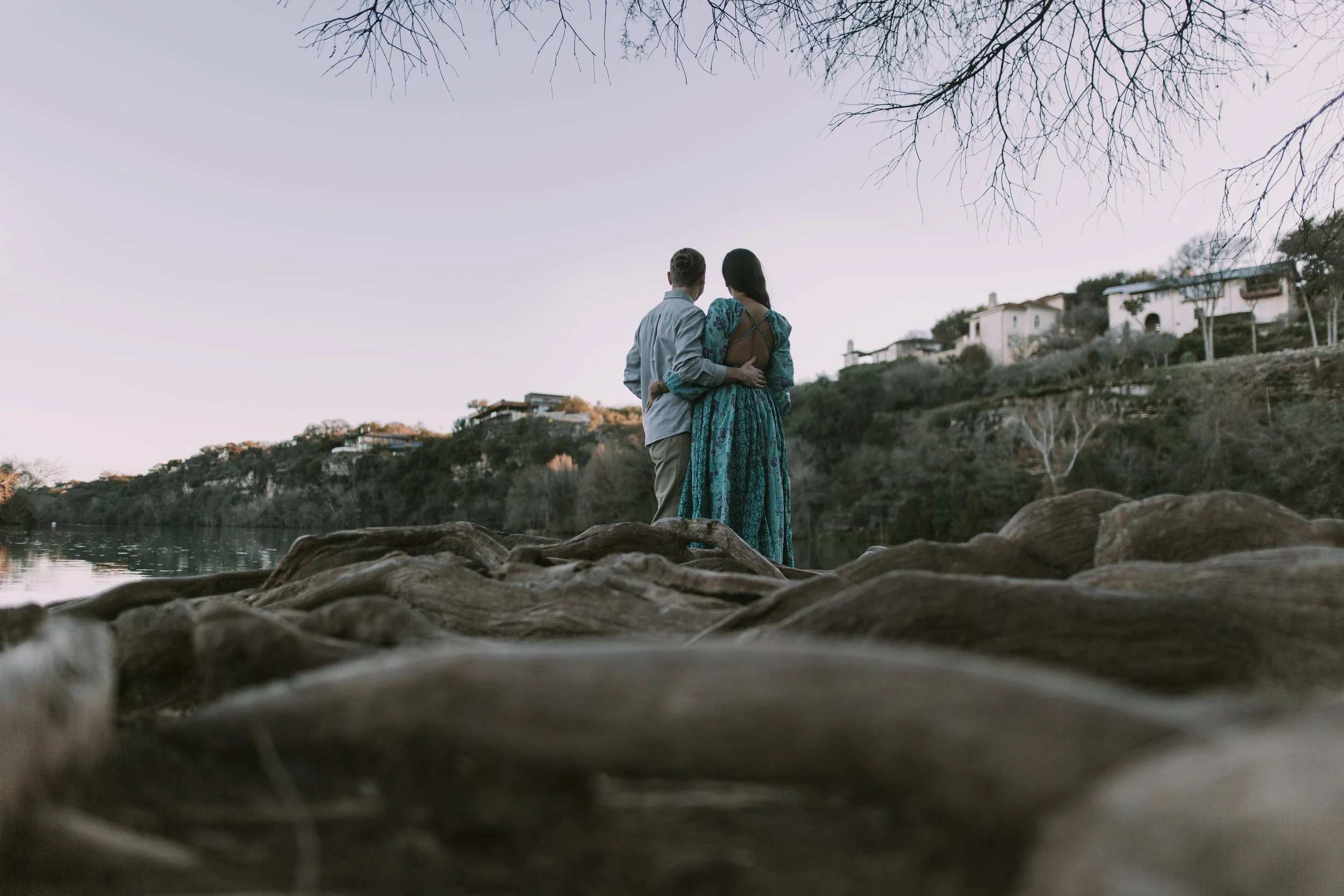 Couple on bald cypress roots above Lake Austin at dusk, hillside houses across the water