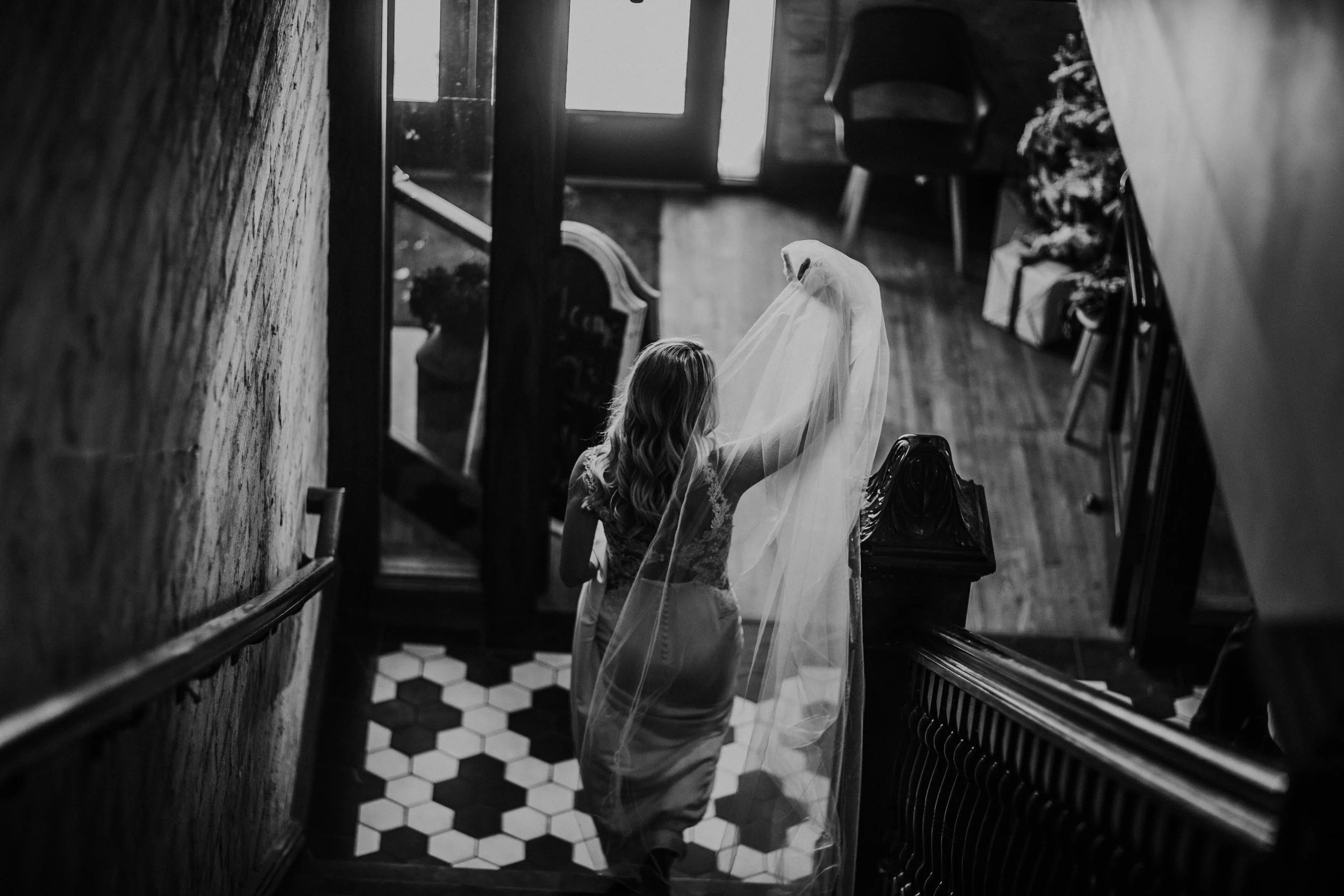 Bride carries her long veil down a staircase over hexagon tile, in black and white