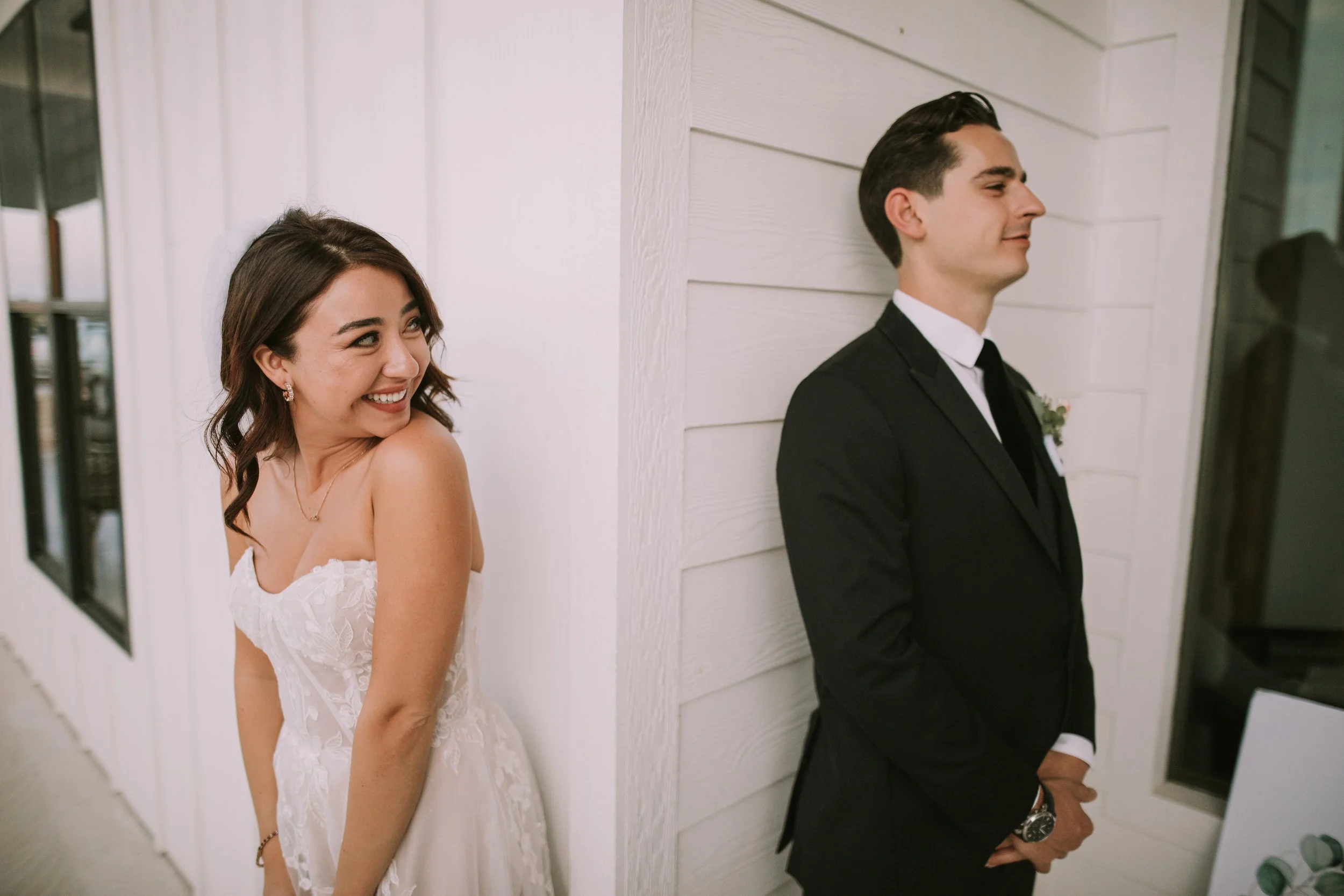 Bride laughs around a white wall corner from her groom in a black tuxedo, moments before their first look