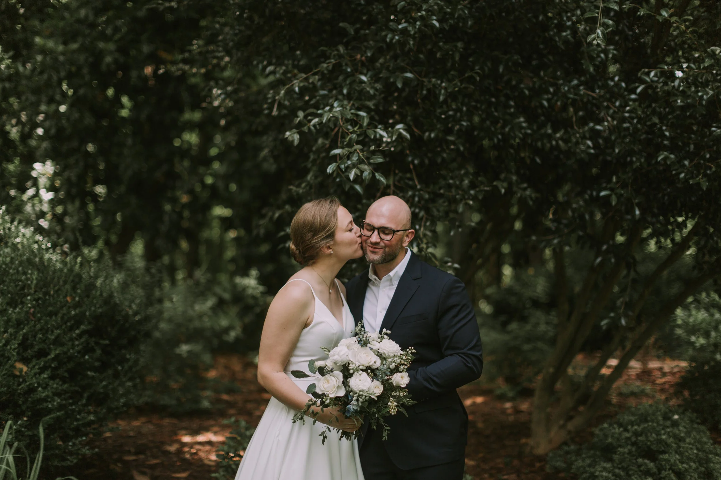 Bride with a white rose bouquet kissing her groom's cheek under garden trees at The Goodyear House in Charlotte