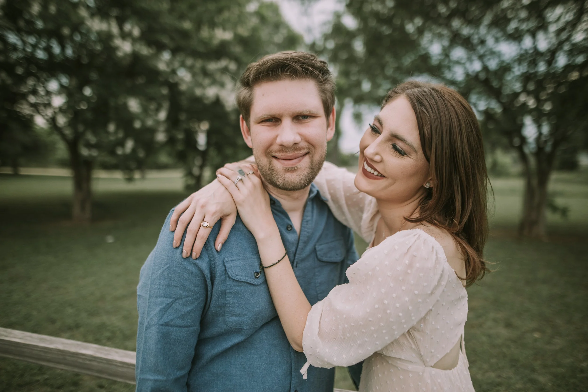 Molly hugging Shane from behind in a tree-lined Austin park, engagement ring catching the soft light