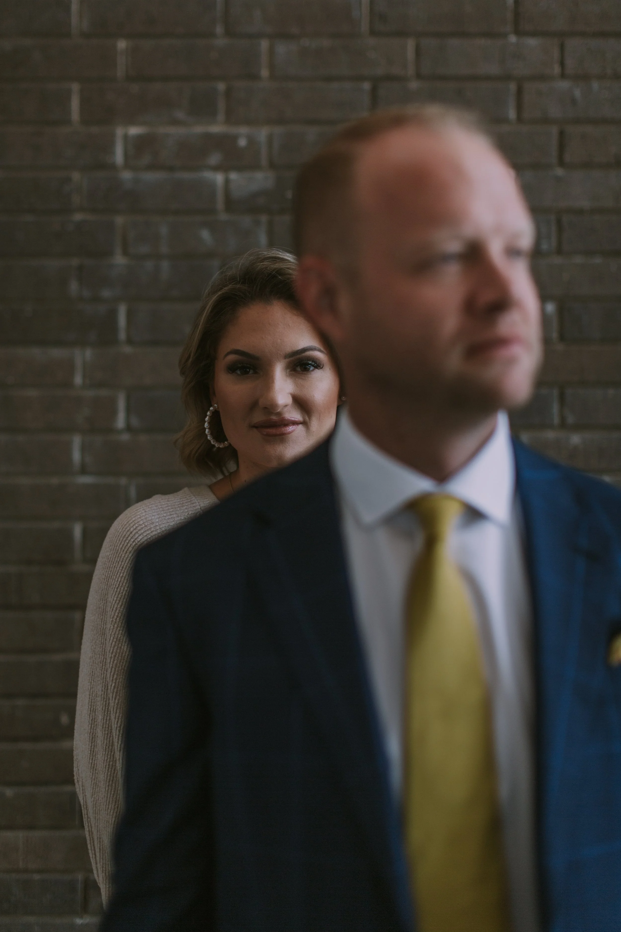 Woman with styled hair in focus behind a man in a navy suit and gold tie, framed against grey brick