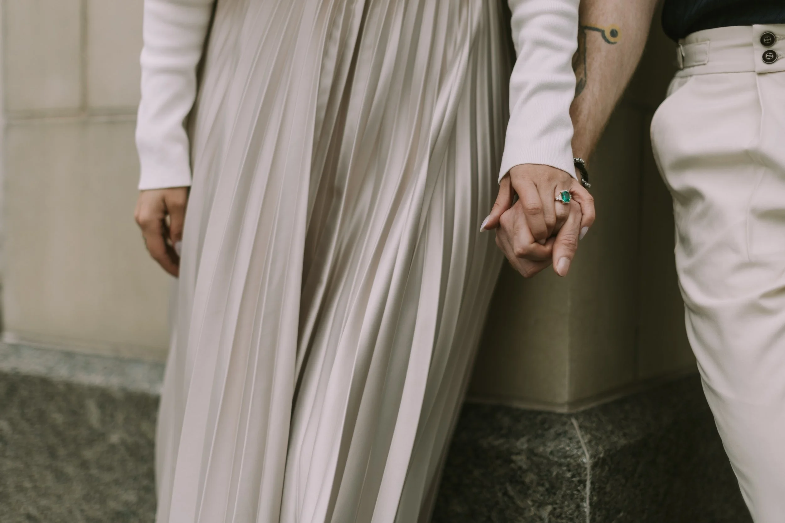 Close-up of interlaced hands with an emerald engagement ring against a pale stone wall in Fort Worth